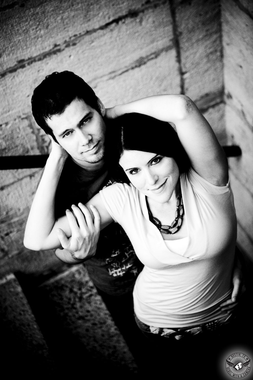 confident, happy girl with dark hair in light shirt and large beaded necklace stands and holds in front of a stoic guy with dark hair and a dark shirt in a stairwell at the Texas Capital in this romantic black and white engagement picture in Austin.